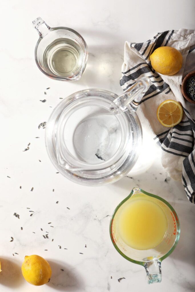 Ingredients to make lavender lemonade in pitchers on marble with lavender buds and lemons