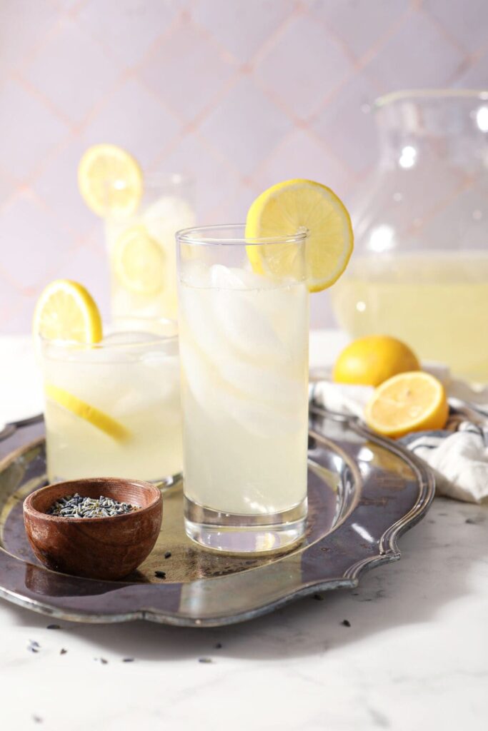 Three glasses of lavender lemonade on a silver serving platter with lavender buds and a pitcher