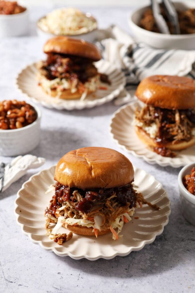 Three plates of barbecue pork sandwiches with slaw and sauce with bowls of baked beans