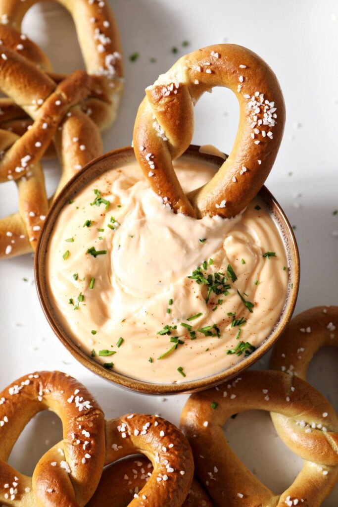 A soft pretzel sits in a bowl of homemade cheese dip garnished with chives