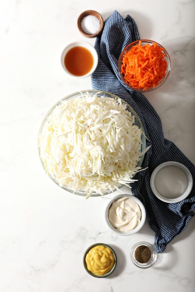 Ingredients to make homemade coleslaw in bowls on a marble countertop with a dark blue towel