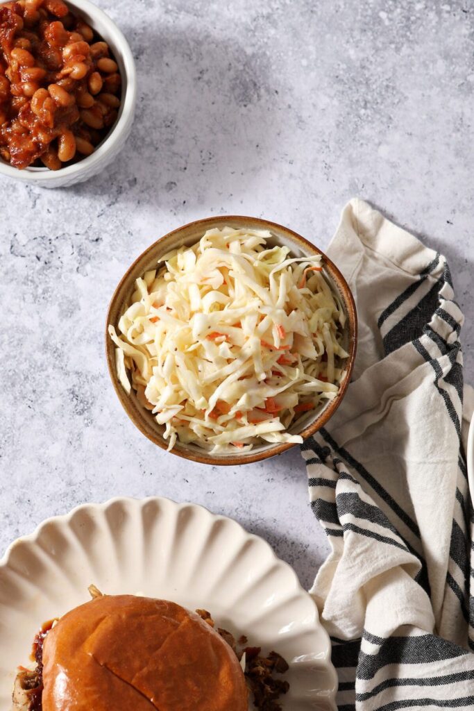 A bowl of coleslaw on a gray textured surface next to a sandwich and a bowl of baked beans