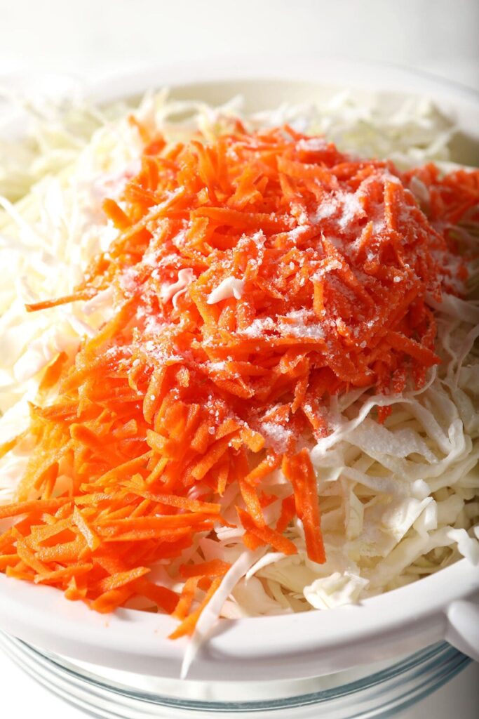 Close up of shredded carrots and cabbage in a white colander seasoned with salt