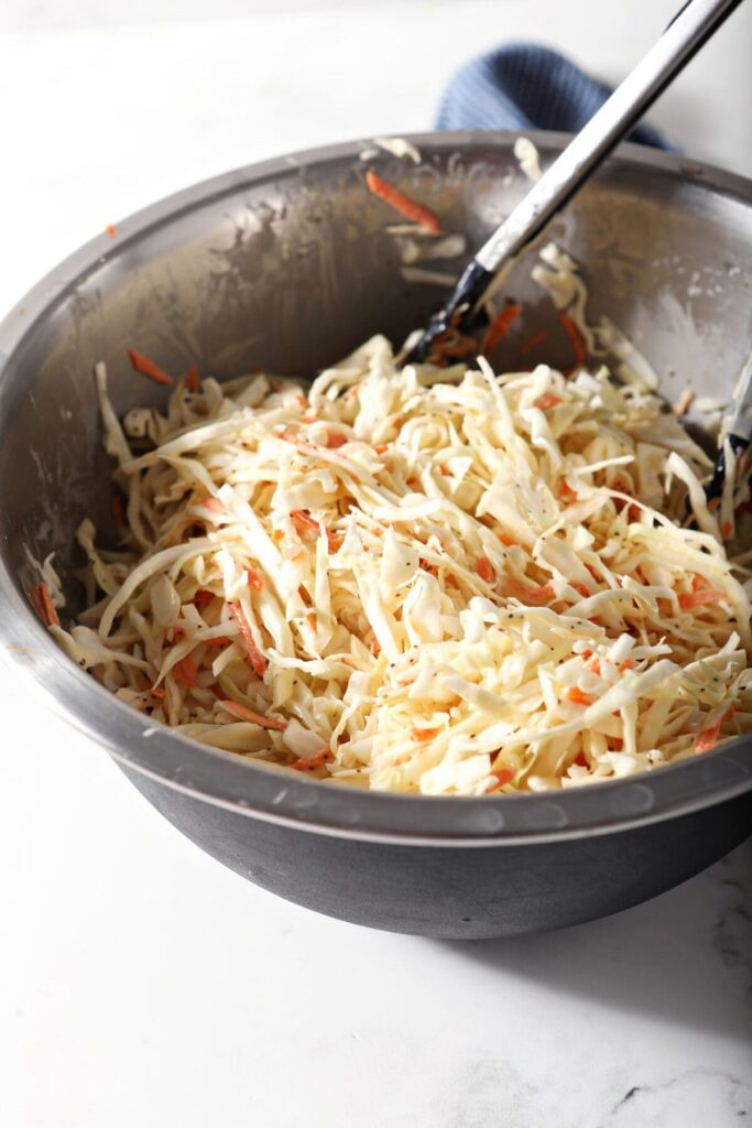 Coleslaw is tossed in a silver and black bowl with tongs