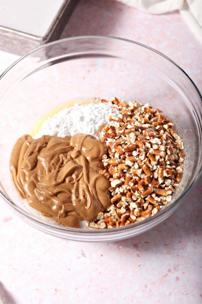 Peanut butter, crushed pretzels, sugar and butter in a bowl on a pink surface