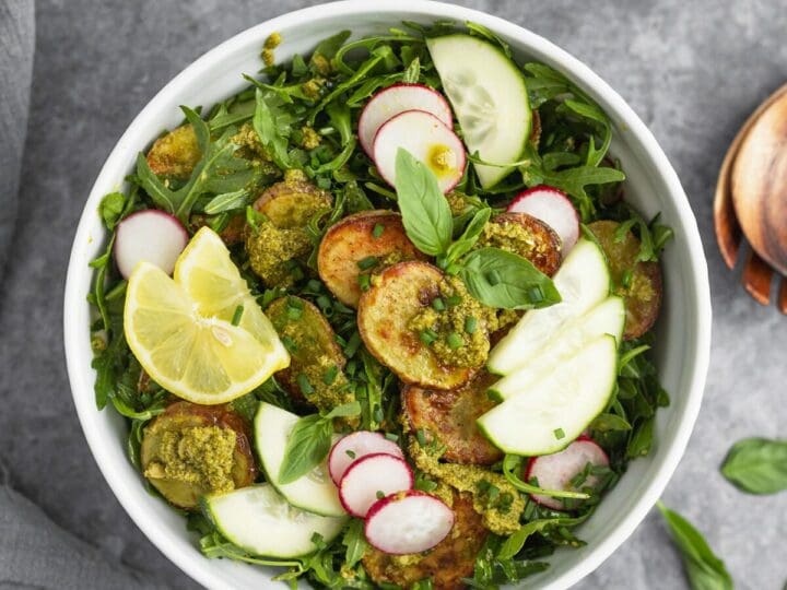 A bowl of roasted potato salad with pesto on a gray table with basil leaves, lemon and a bowl of pesto