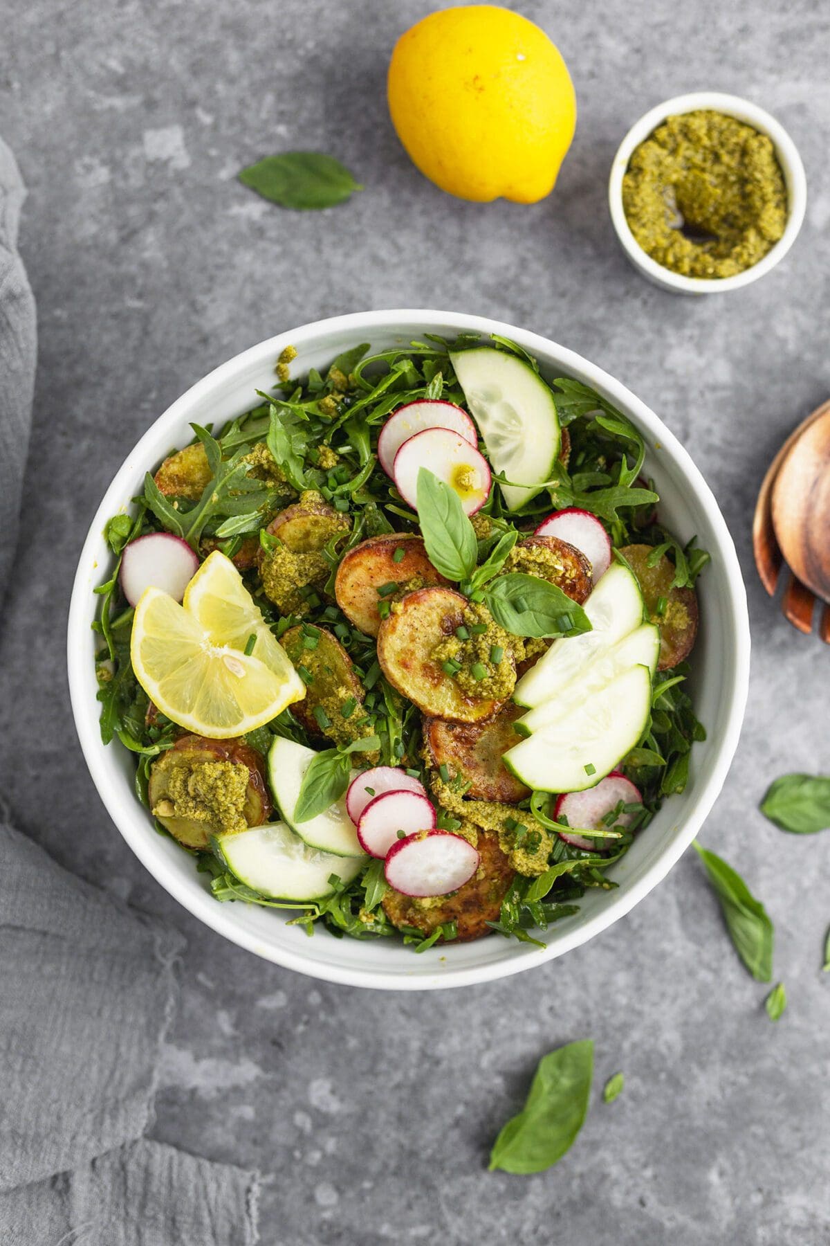 A bowl of roasted potato salad with pesto on a gray table with basil leaves, lemon and a bowl of pesto