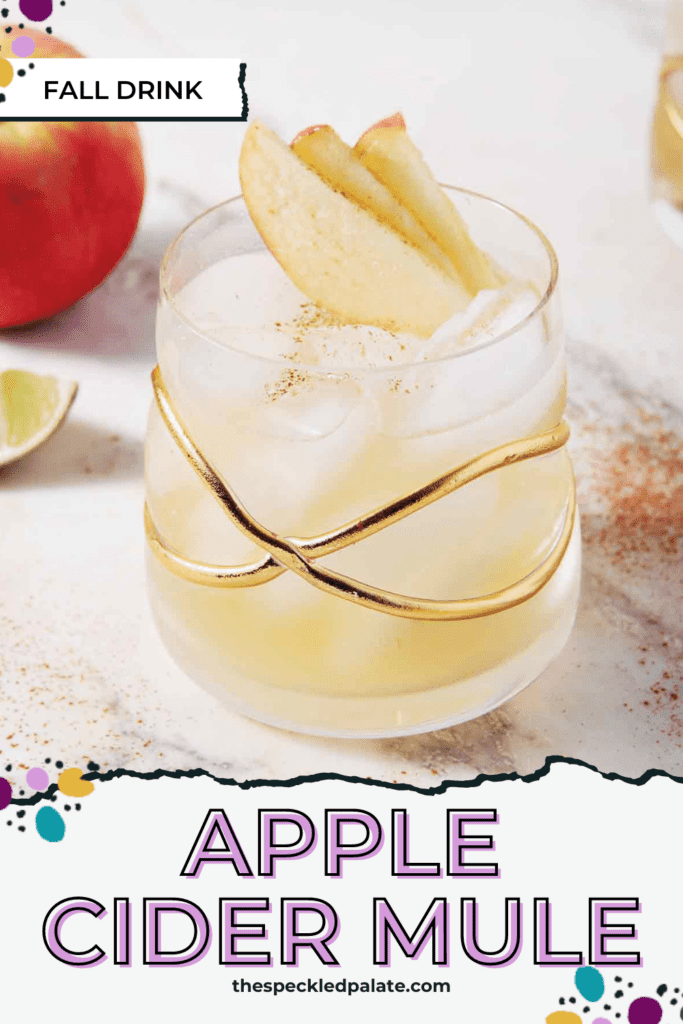 An Apple Cider Moscow Mule sit on a marble background, surrounded by apples, lime slices and cinnamon with the text Apple Cider Mule