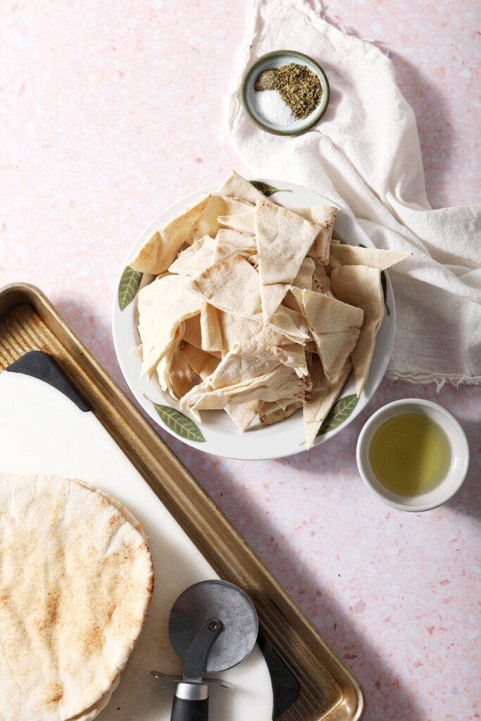 Sliced pita triangles in a bowl next to a cutting board and bowls of olive oil and a seasoning blend