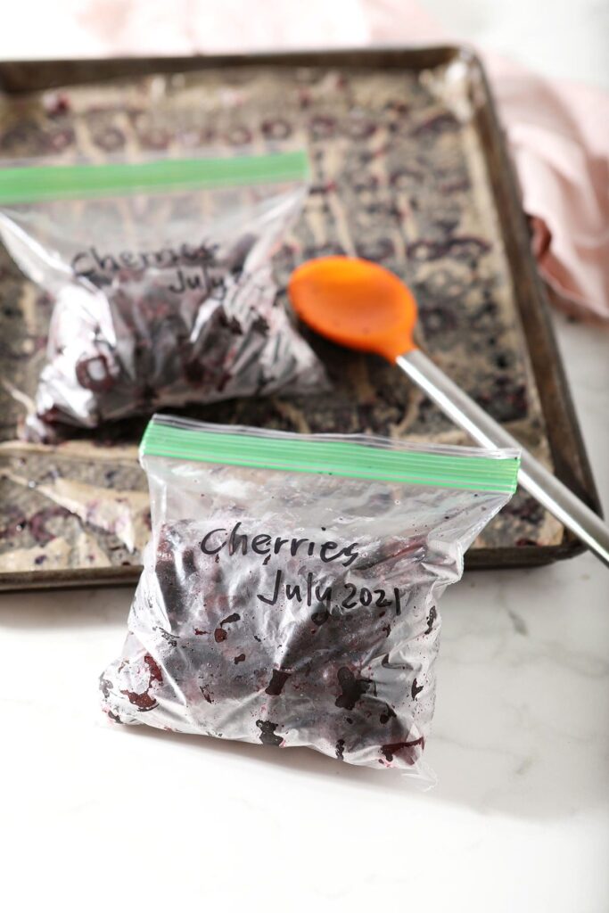 Two plastic freezer bags of frozen cherries next to a sheet pan with an orange spoon