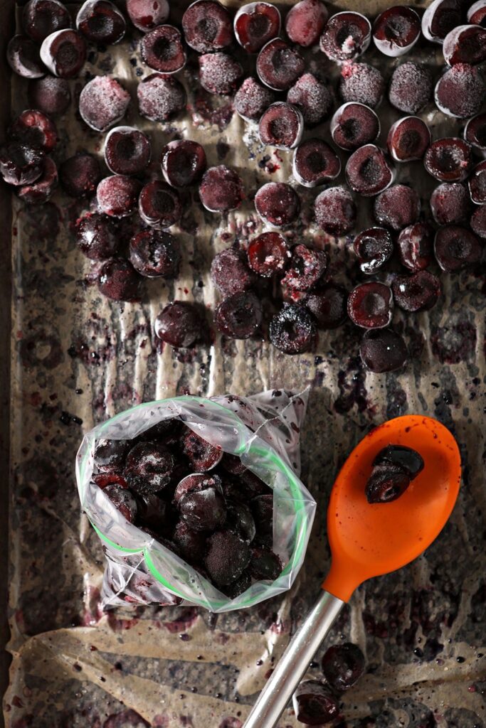 Frozen cherries on a sheet pan with a plastic bag and an orange-tipped spoon before going into the deep freeze