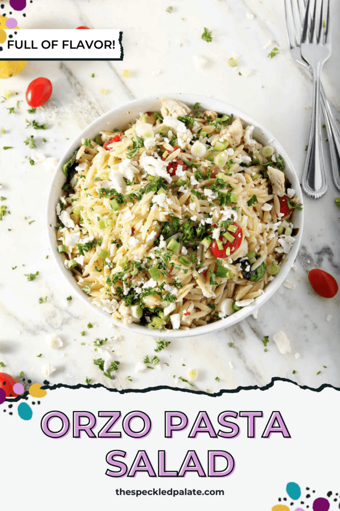 Overhead of a bowl of Greek Orzo Pasta Salad on marble, surrounded by tomatoes, chopped parsley, lemon and red onion with the text orzo pasta salad