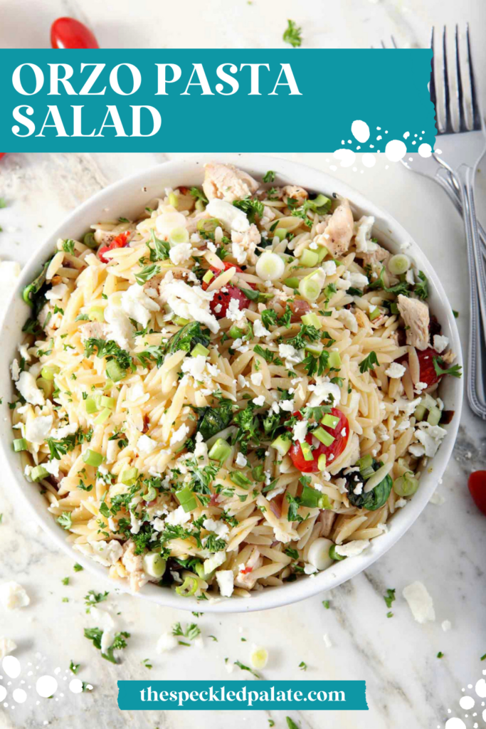 Close up overhead of a bowl of Greek Orzo Pasta Salad on marble, surrounded by tomatoes, chopped parsley, lemon and red onion with the text orzo pasta salad