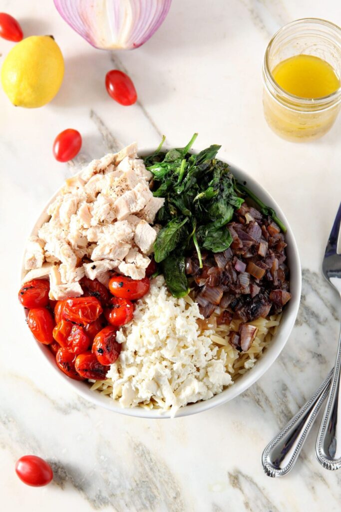 A bowl of ingredients, including charred tomatoes, chicken, wilted spinach, red onion and feta cheese in a bowl on marble