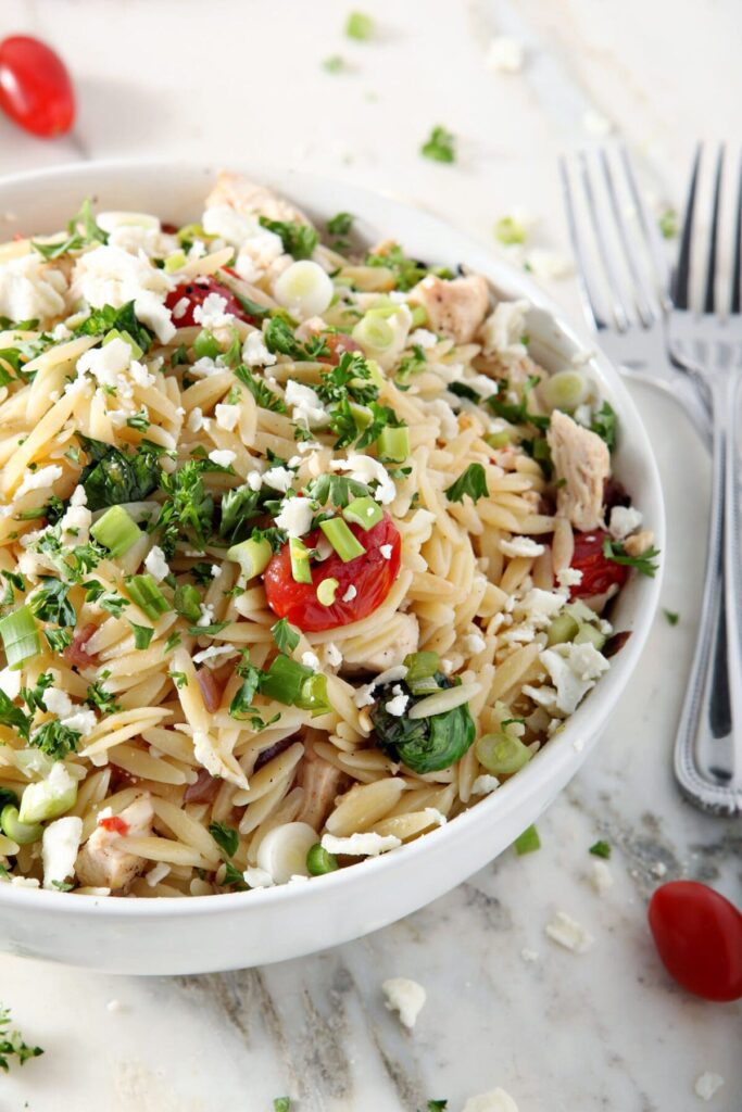 Close up side view of a white bowl of orzo pasta salad with chicken, garnished with green onions and fresh parsley