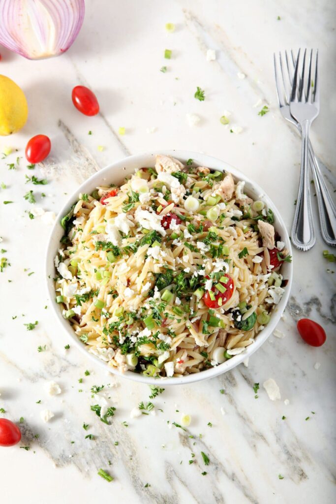 Overhead of a bowl of Greek Orzo Pasta Salad on marble, surrounded by tomatoes, chopped parsley, lemon and red onion