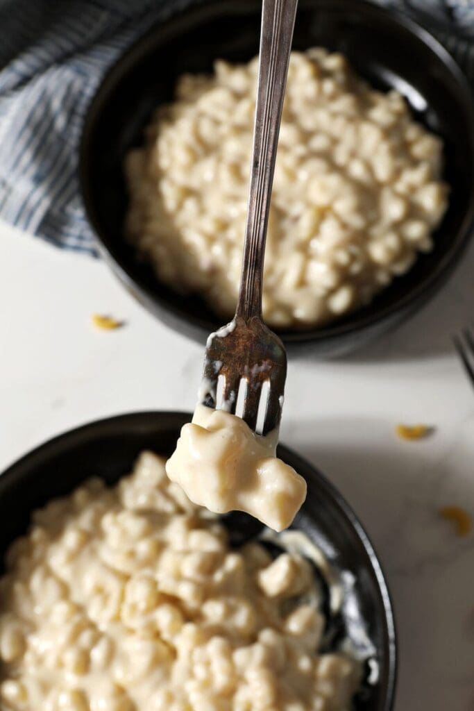 A fork holds a bite of three cheese Mac and cheese above two black bowls holding more macaroni and cheese