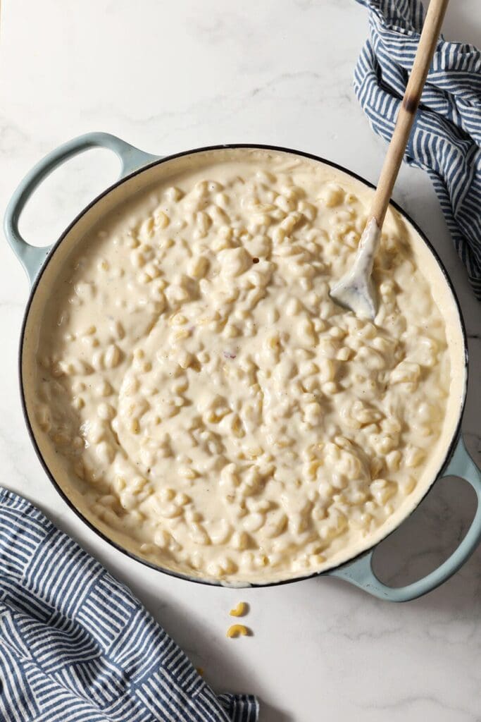 Homemade macaroni and cheese in a braiser pan with a wooden spoon inside it on marble next to patterned blue napkins