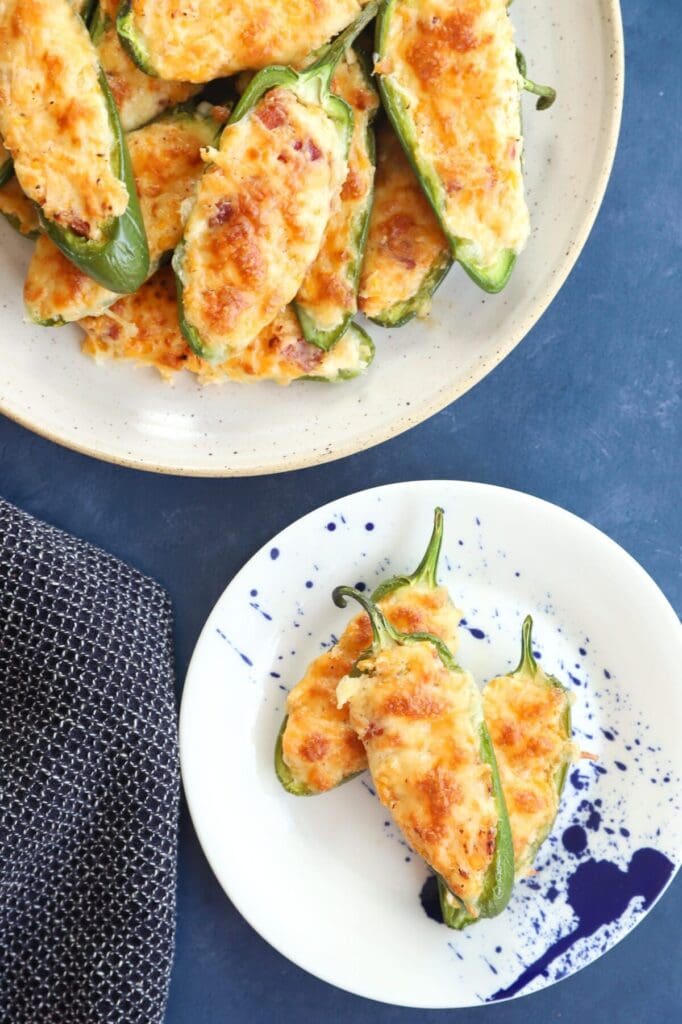 A small plate holds three jalapeno poppers next to a larger platter of poppers on a blue surface