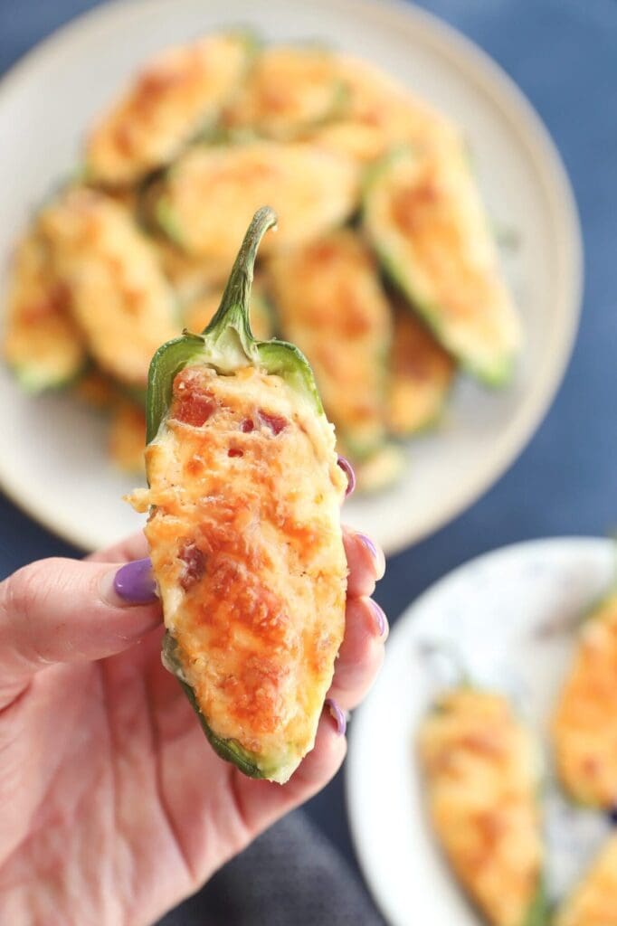 A woman holds a golden brown cheesy jalapeño popper in hand above a large serving platter