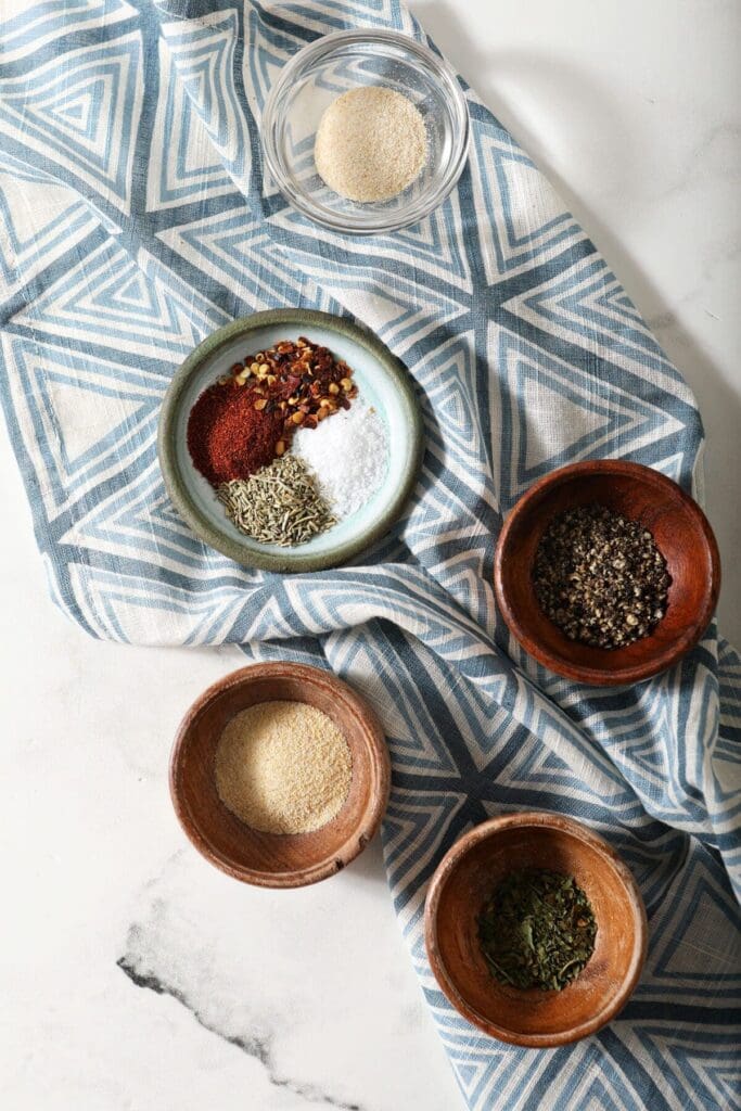 Ingredients to make a steak seasoning blend in bowls on a marble countertop with a blue patterned towel