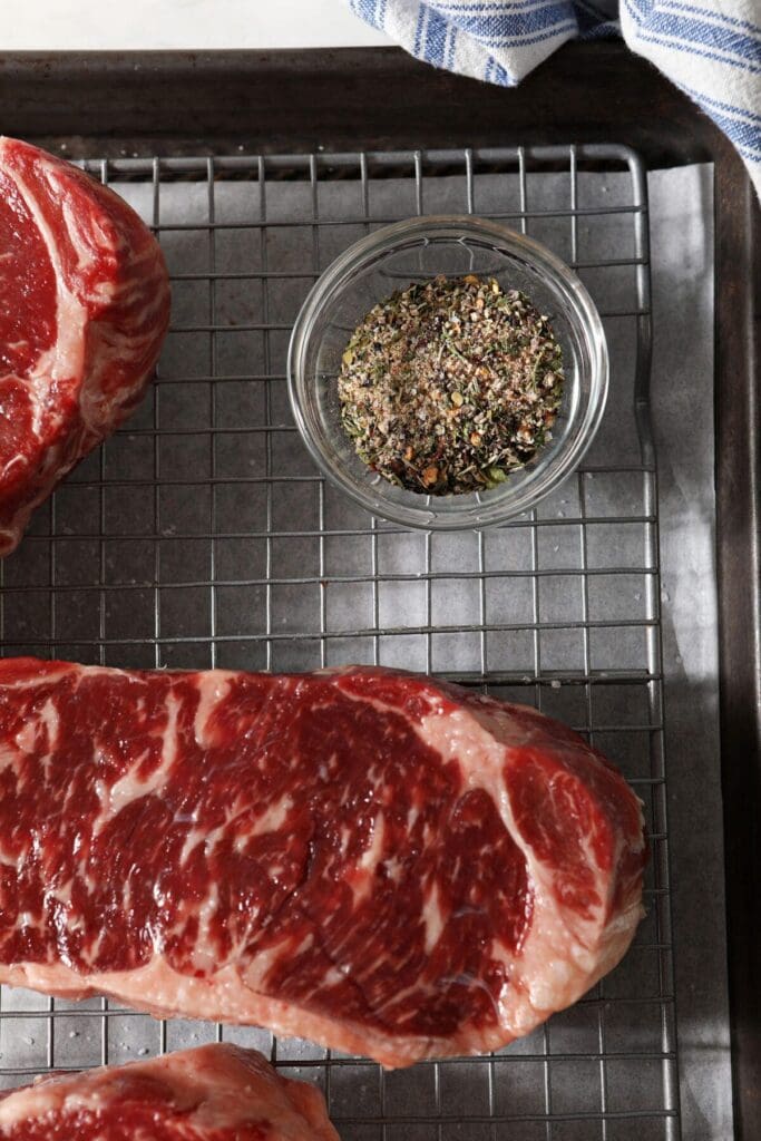 Steak seasoning blend in a small glass bowl next to steaks on a wire rack