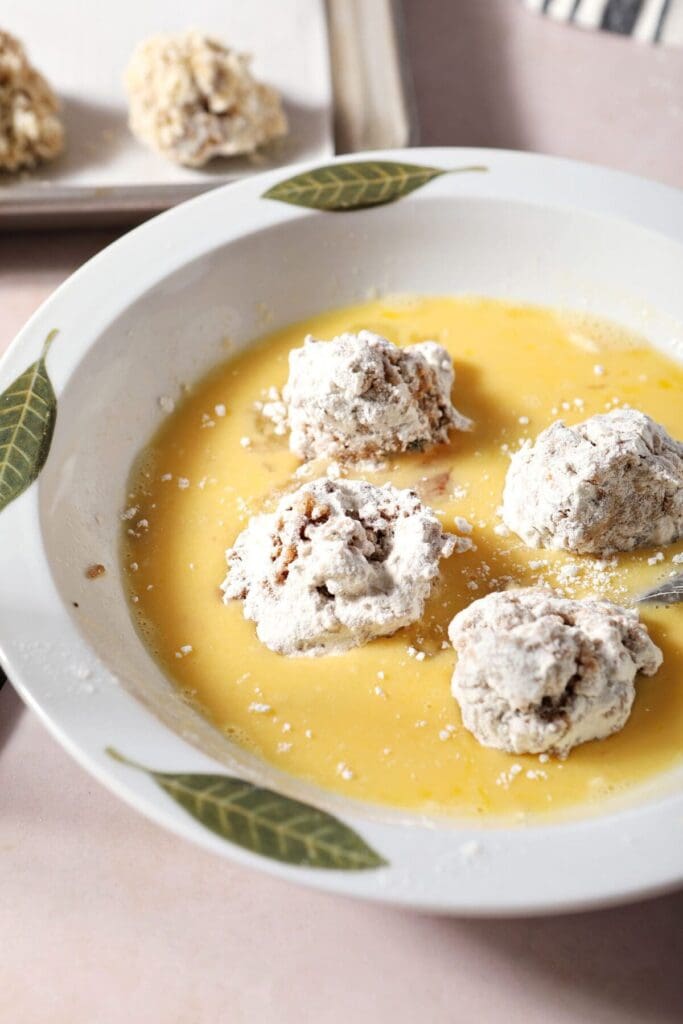 Four boudin balls coated in flour in an egg wash mixture in a bowl