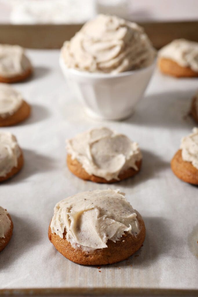 Frosted pumpkin cookies line a pan with a bowl of brown butter frosting in the background