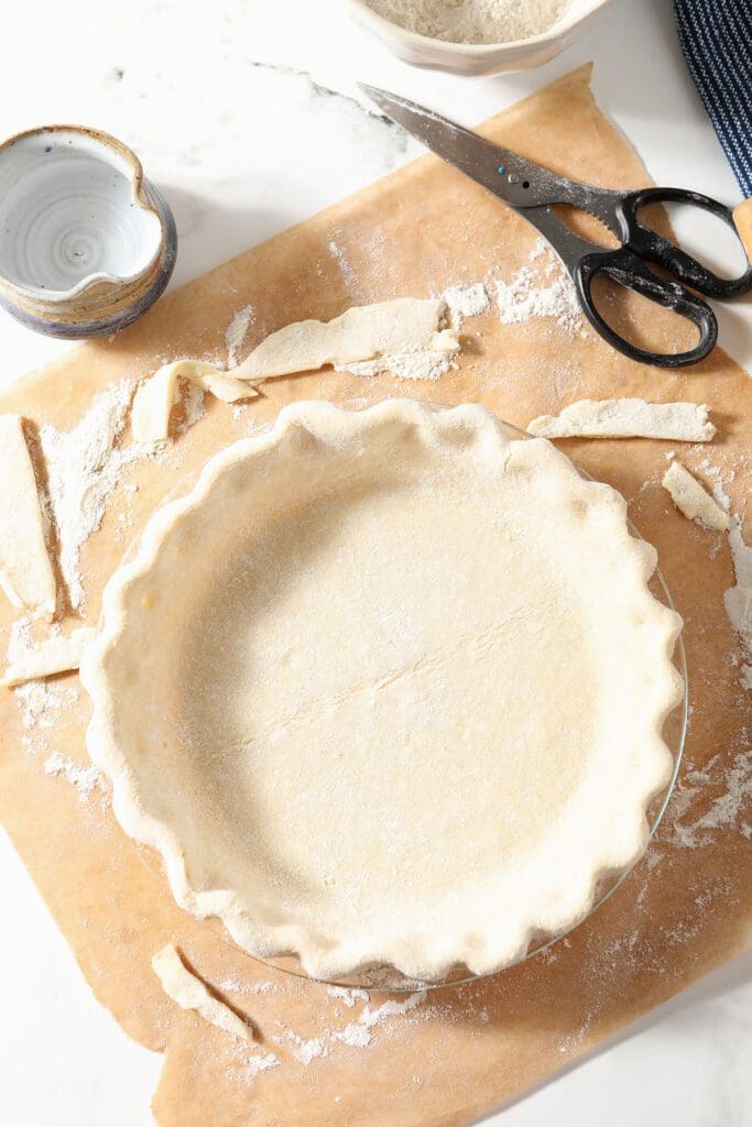 An unbaked pie crust on a piece of brown parchment paper surrounded by scraps, scissors and a bowl of vodka and a bowl of flour