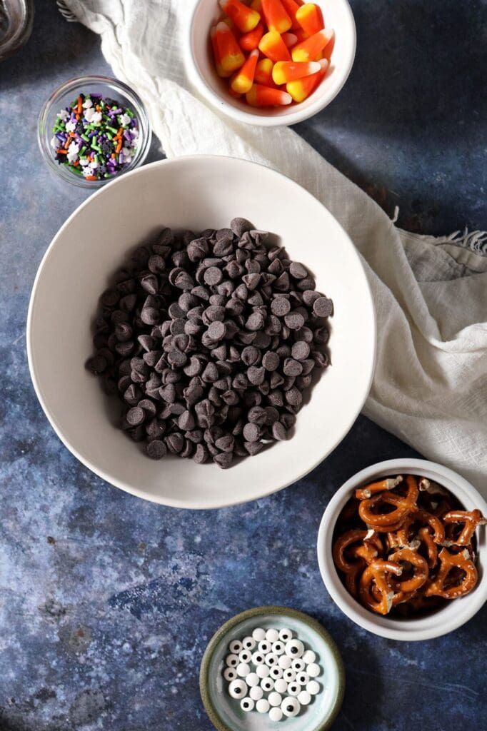 ingredients to make a chocolate bark with pretzels and halloween candies in bowls