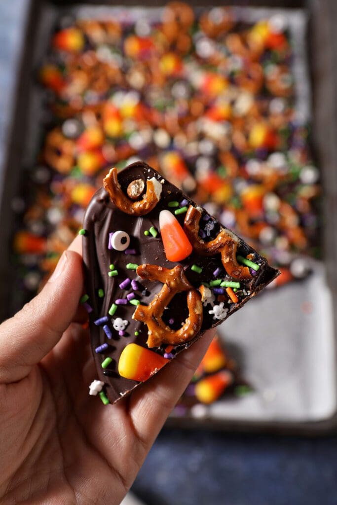 a hand holds a slice of halloween bark with pretzels and candy corn above a sheet pan