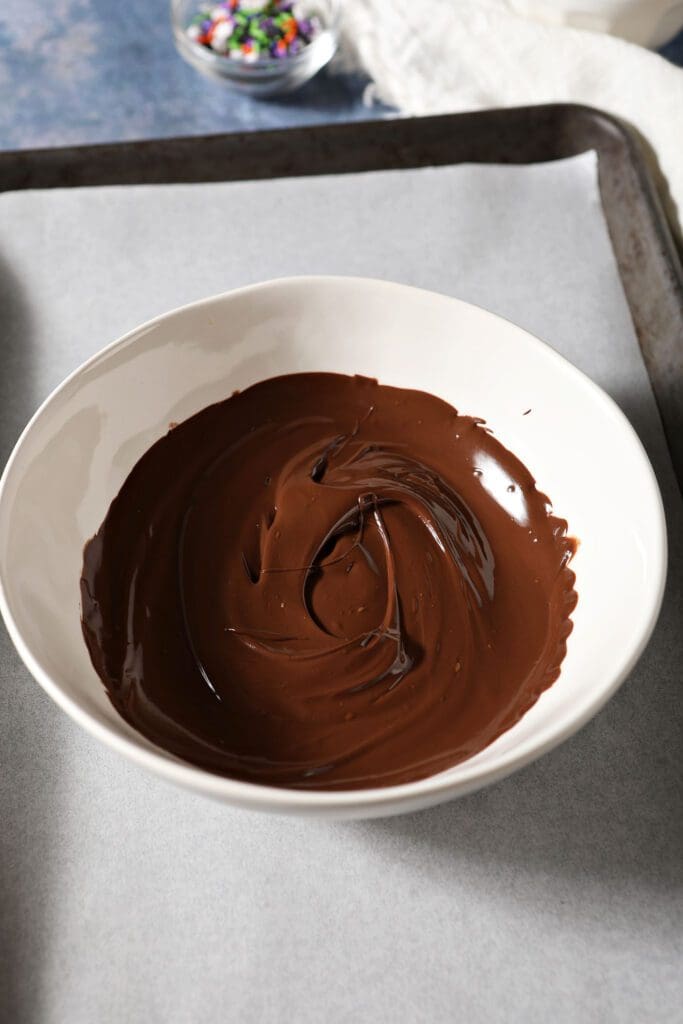 a white bowl holds melted chocolate on a parchment-lined jelly roll pan