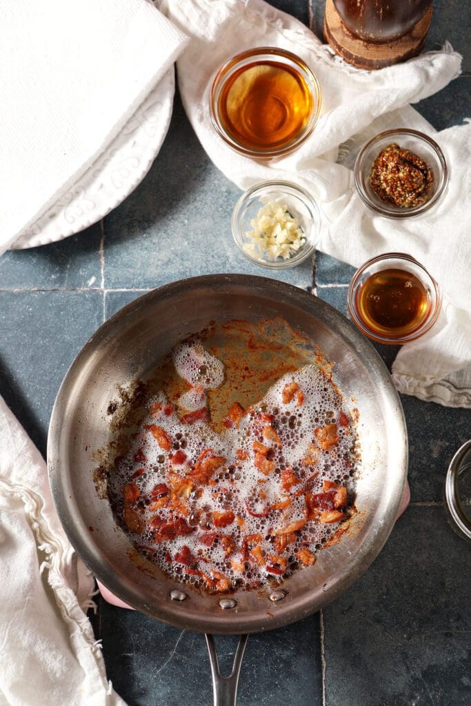 Bacon sizzles in a pan while the rest of the vinaigrette ingredients sit next to it in bowls on a tiled blue surface