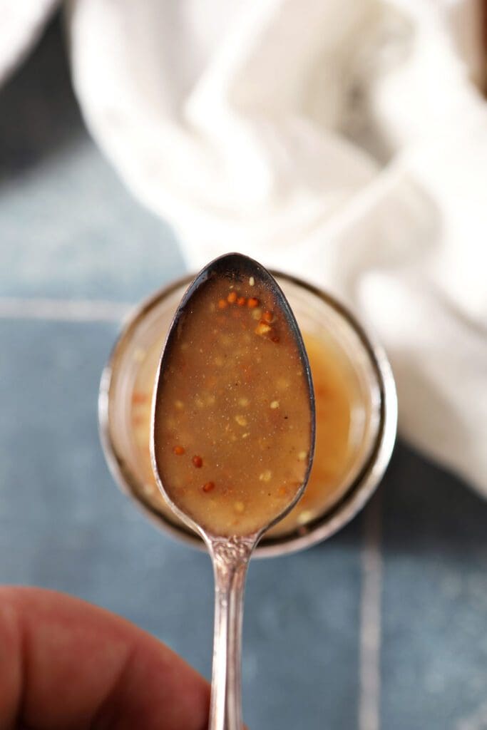 A spoon holds a serving of bacon vinaigrette above a jar of the dressing next to a white towel on a blue tiled surface