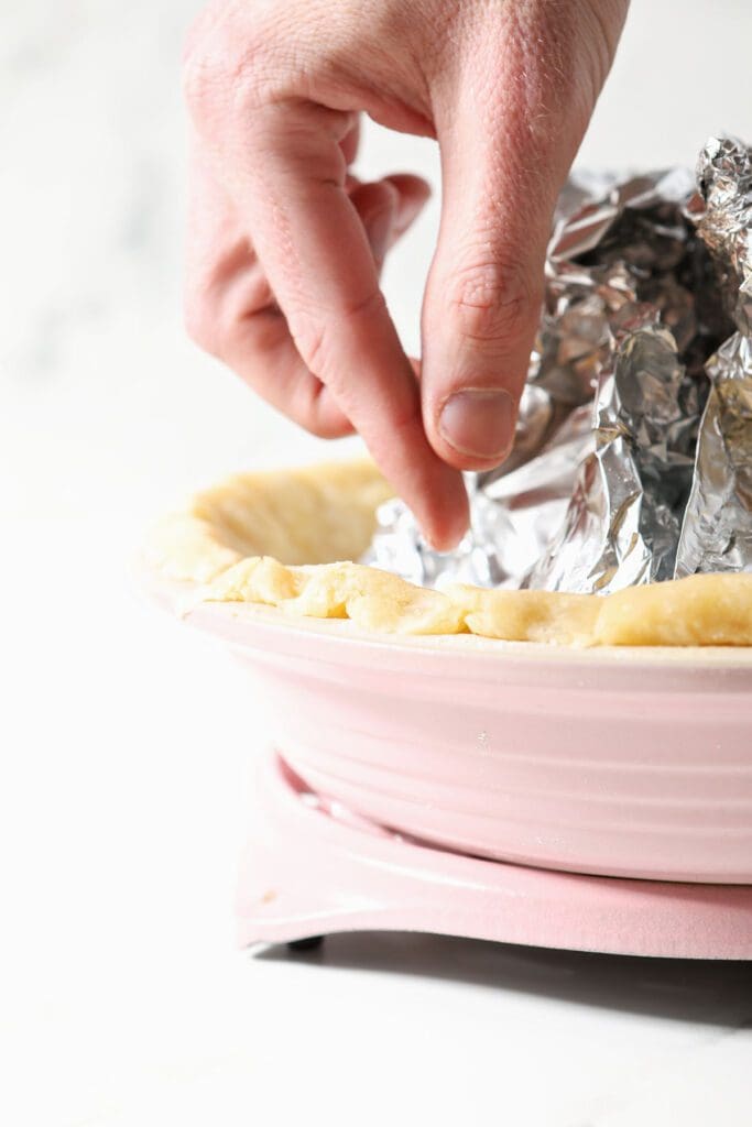 A hand lifts foil out of a blind baked pie crust