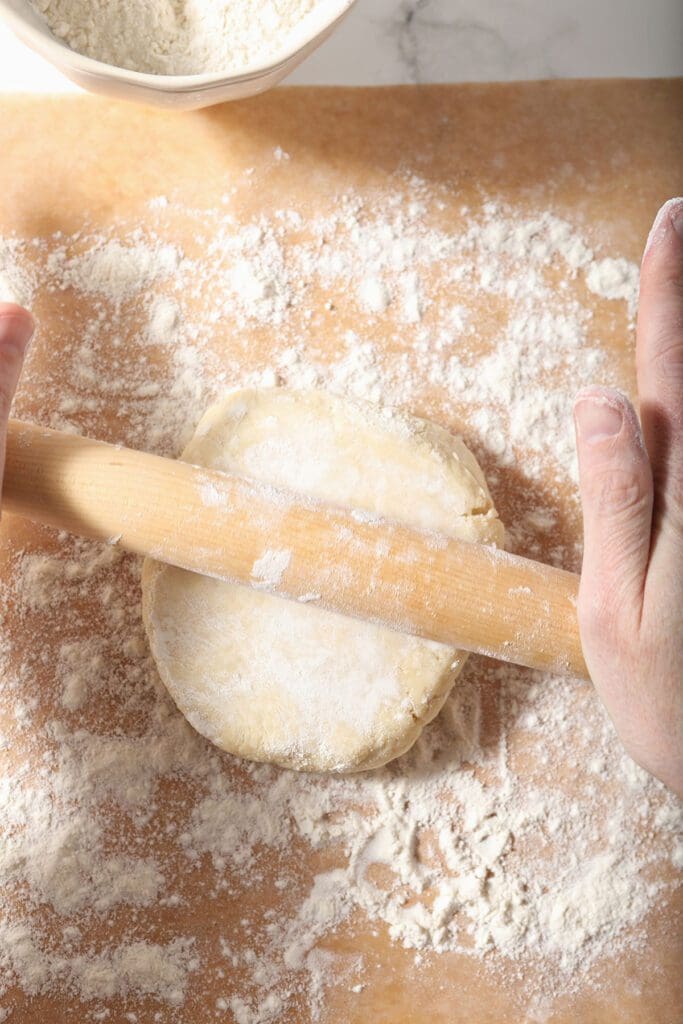 A round of pie crust is rolled out with a wooden rolling pin on a floured sheet of brown parchment