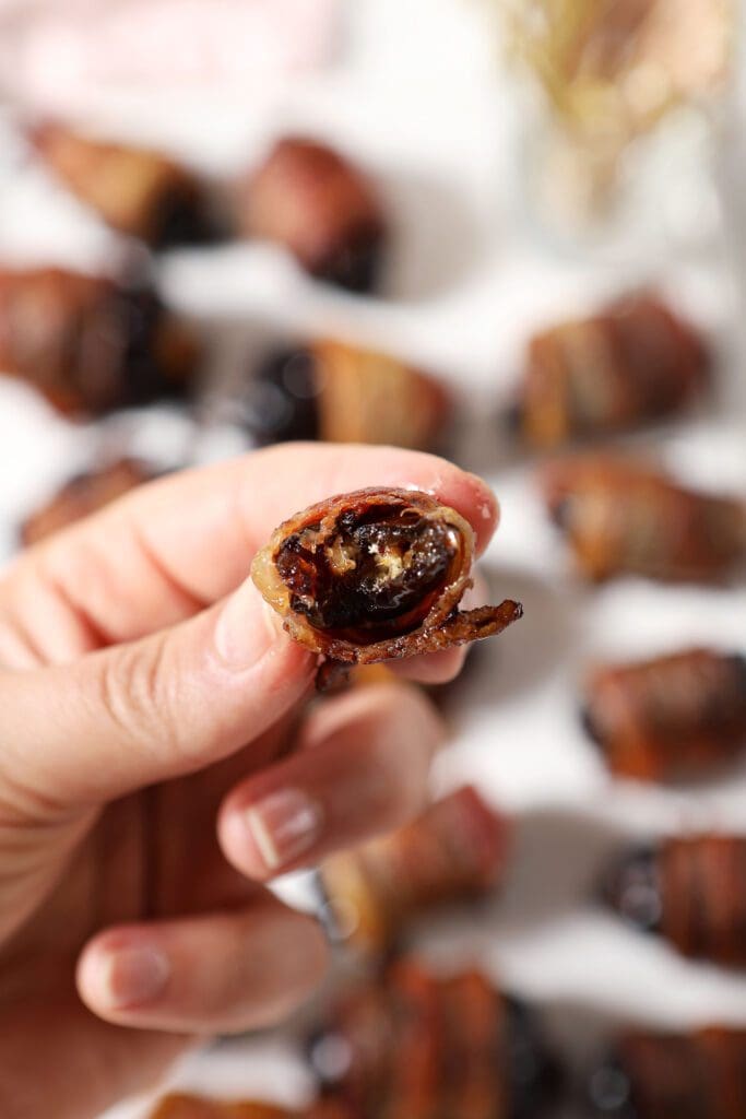 A hand holds a bitten-into bacon wrapped date above a platter of more dates