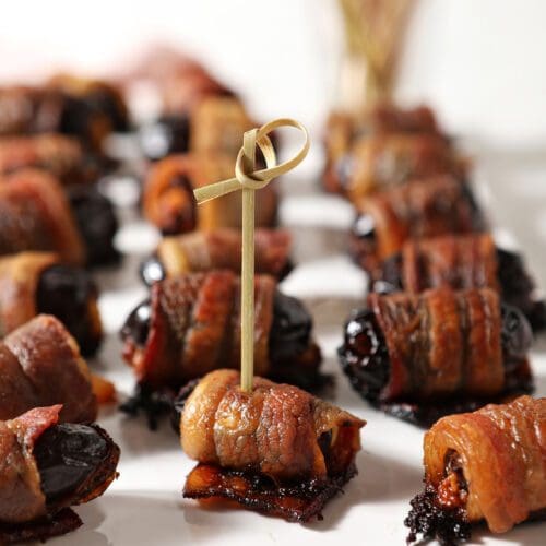 A wooden toothpick skewers a bacon wrapped date on a platter of stuffed dates