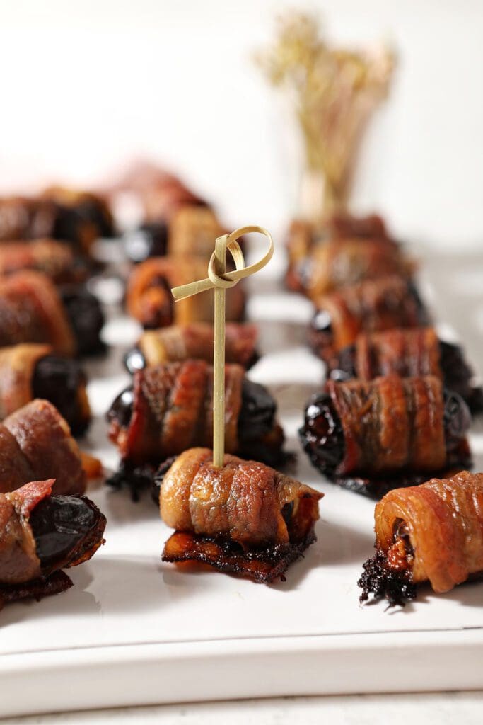 A wooden toothpick skewers a bacon wrapped date on a platter of stuffed dates