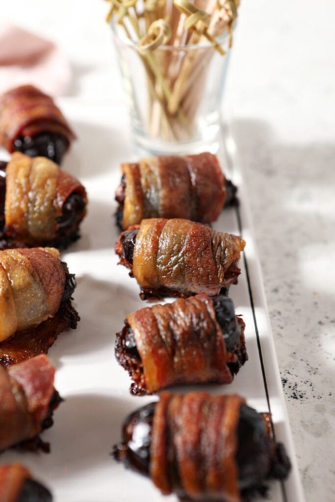 A white platter of bacon wrapped dates with a glass container of wooden toothpicks