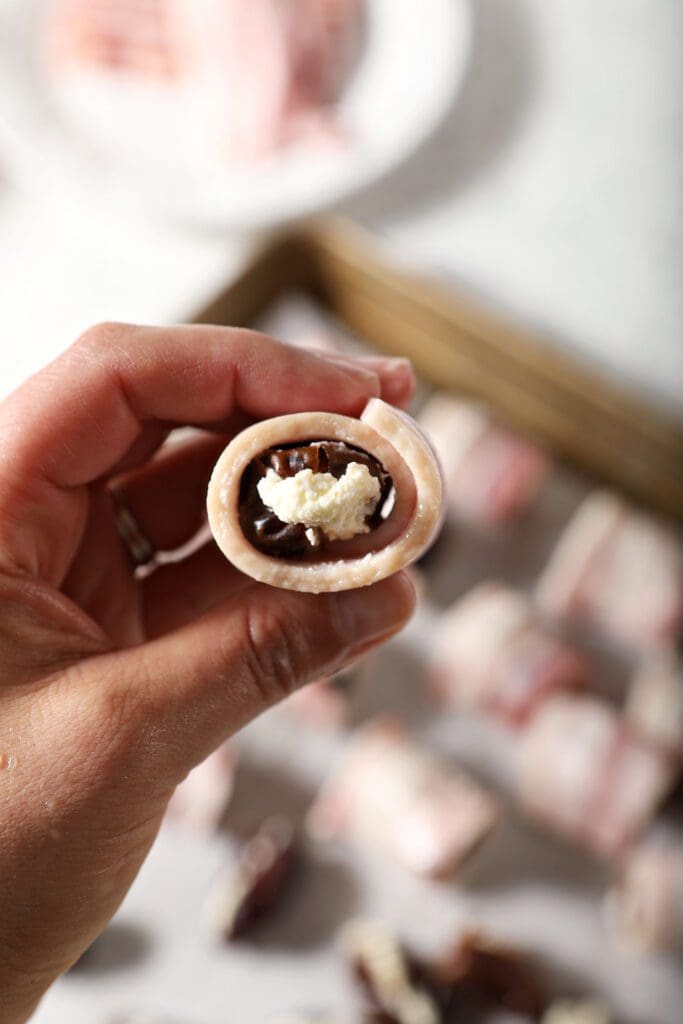 A stuffed date rolled in a piece of bacon in a hand above a sheet pan of more bacon wrapped dates before baking