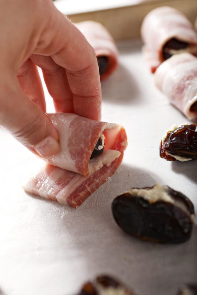 A hand rolls a feta stuffed date in a strip of bacon