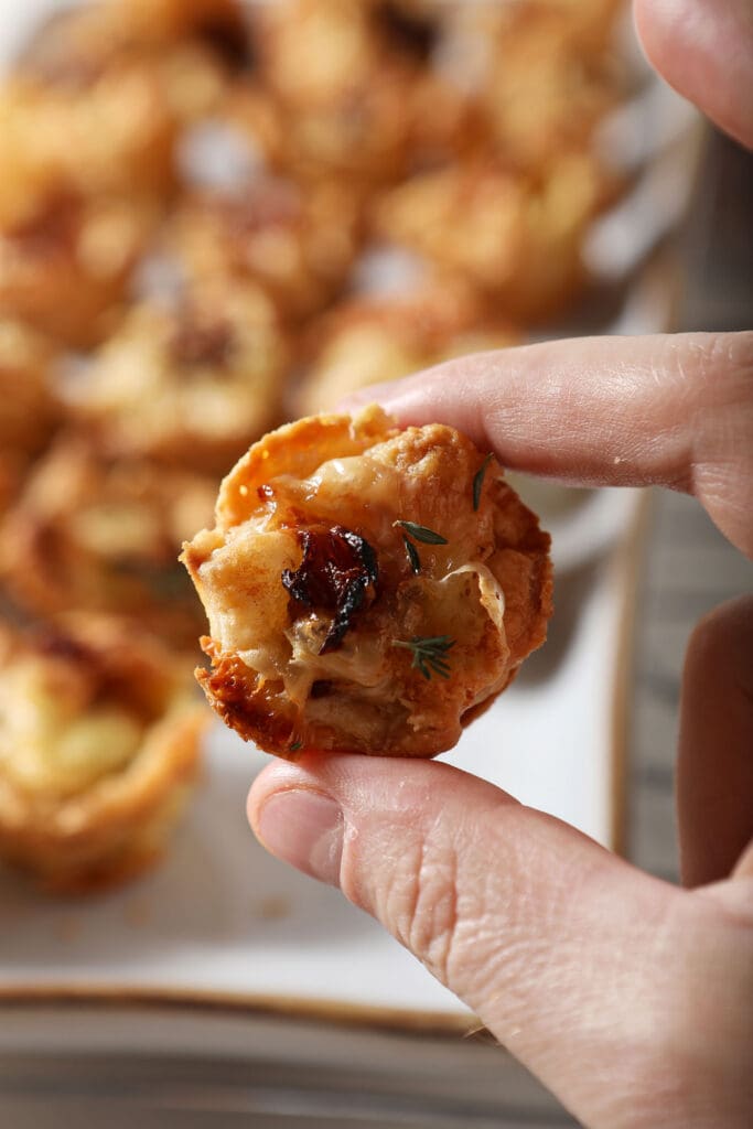 A hand holds a baked brie bite in front of a white platter of more brie bites