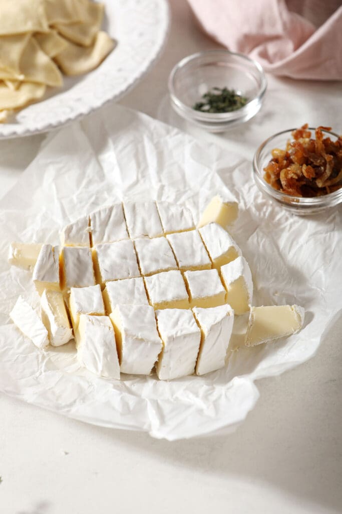 A sliced wheel of brie on a white surface surrounded by bowls of other ingredients