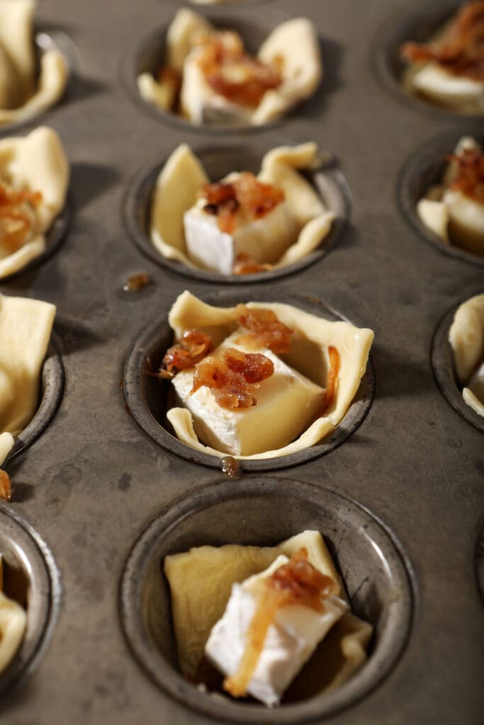 Caramelized onion on top of brie cheese slices in puff pastry in a muffin tin before baking