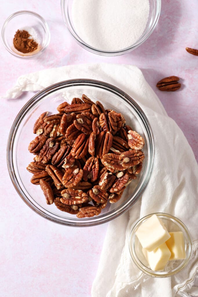 A bowl of pecans next to other ingredients in bowls on a pink surface to make glazed pecans