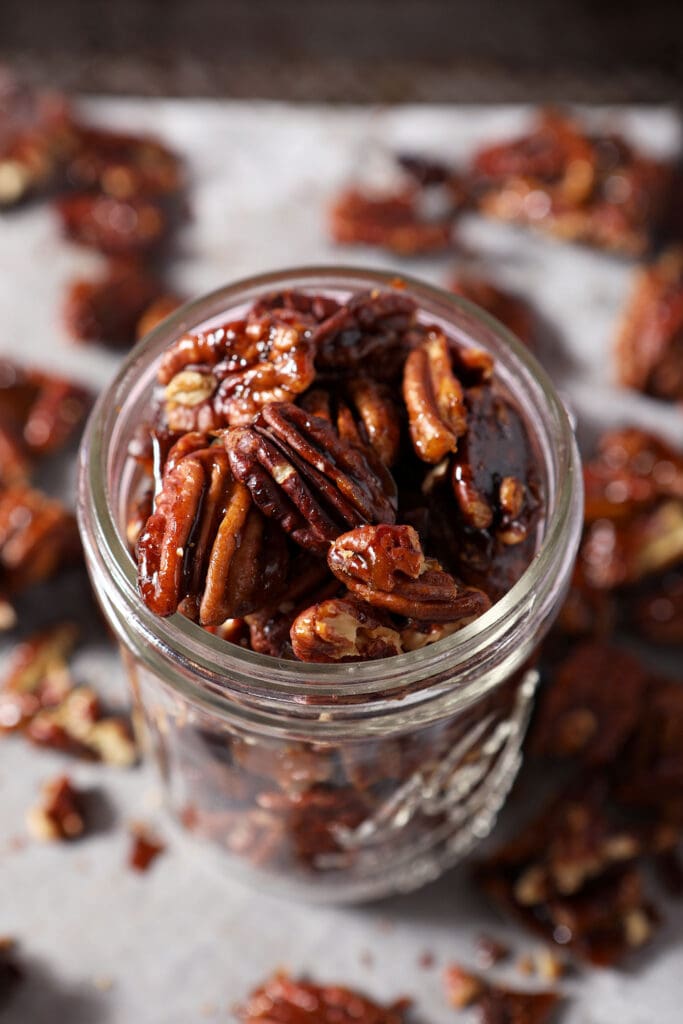 A glass jar of glazed pecans on top of a sheet pan with more pecans