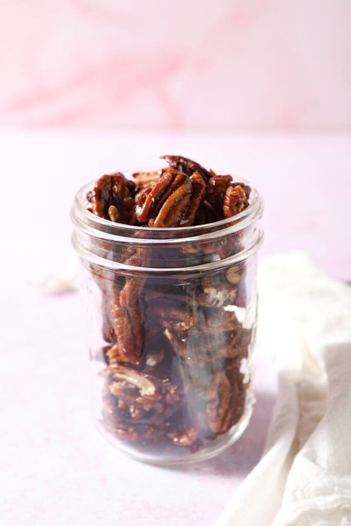 A mason jar of glazed pecans on a pink surface next to a white linen