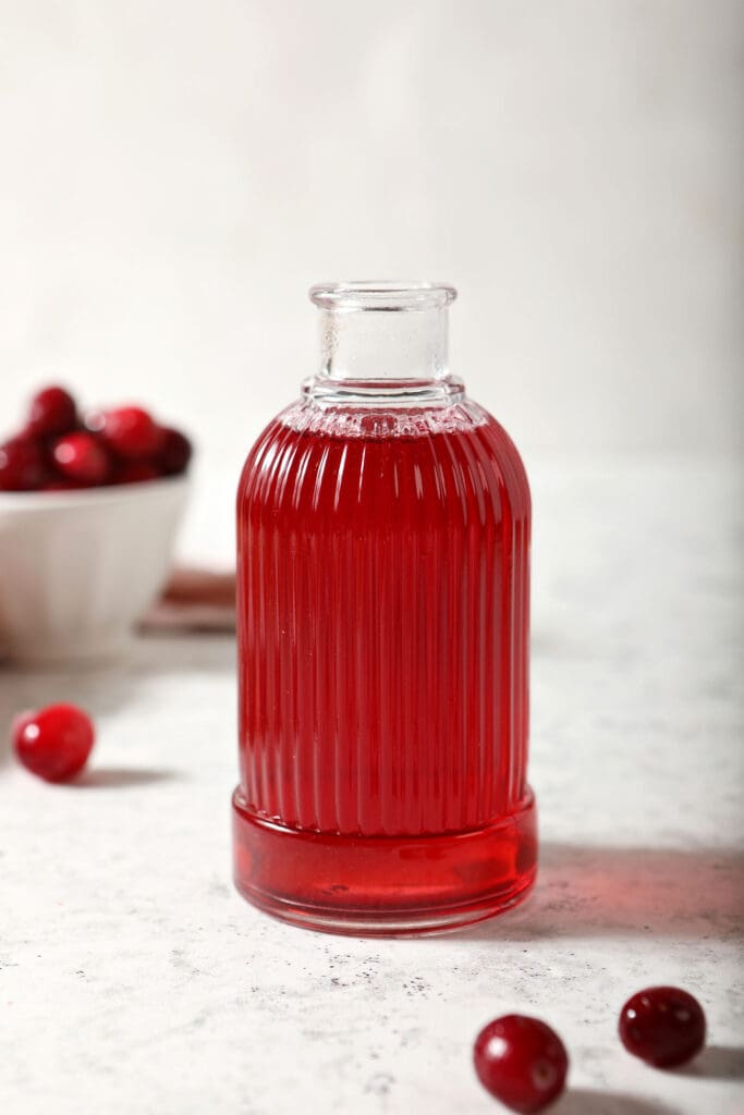 A ribbed glass jar of cranberry simple syrup with cranberries around it on marble