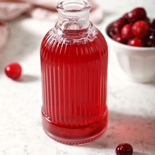 A ribbed glass jar of cranberry simple syrup with a bowl of cranberries and a pink linen on marble